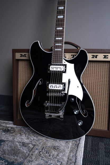 Black Regata LTD electric guitar with a glossy finish, featuring a white pickguard, chrome hardware, and a dark fretboard, and Mosby Tremolo. In front of magnatone  guitar amp.