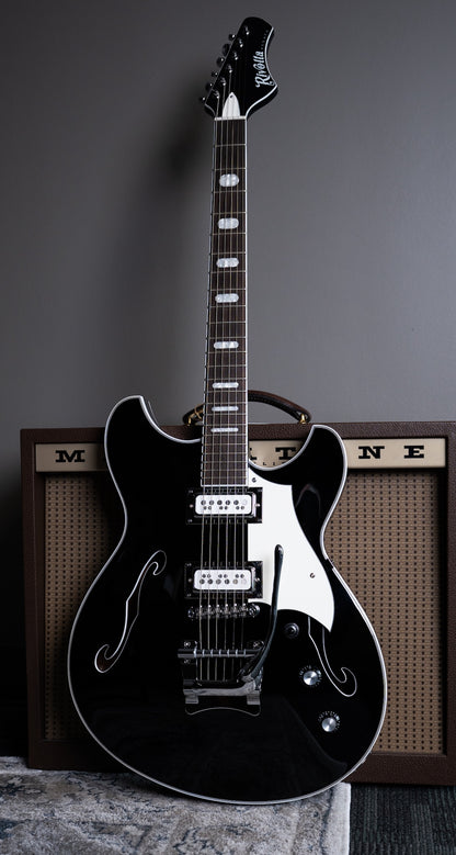 A black Regata LTD semi hollow body guitar with chrome hardware, and a Mosby Tremolo. Standing upright in front of a Magnatone guitar amplifier.